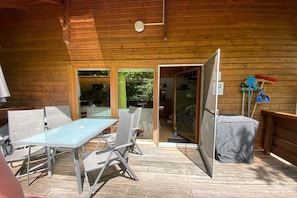 An outdoor wooden deck with a glass door leading to a covered area, a table with chairs, and a covered trash bin. - Holiday home in Edersee with 2 bedrooms (Vöhl \/ Asel)