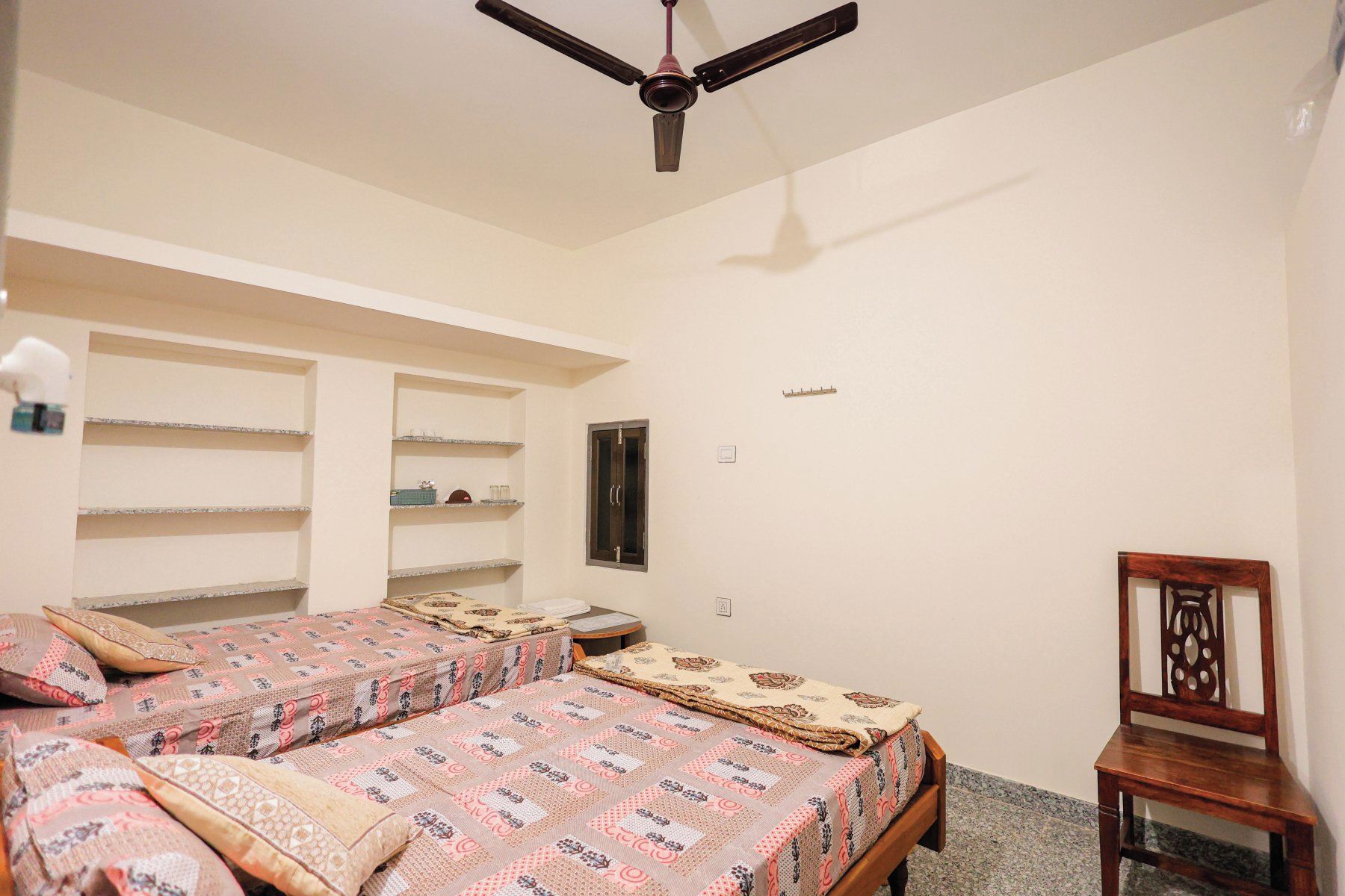 Una habitación con dos camas, un ventilador de techo, una silla de madera y una mesita.