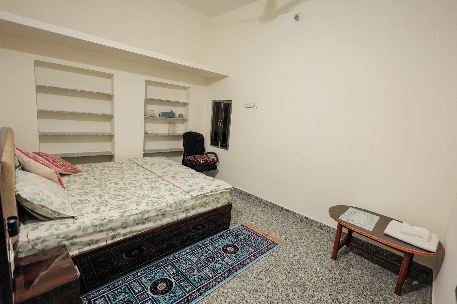 A bedroom with a bed, a chair, a small table, and built-in shelves.