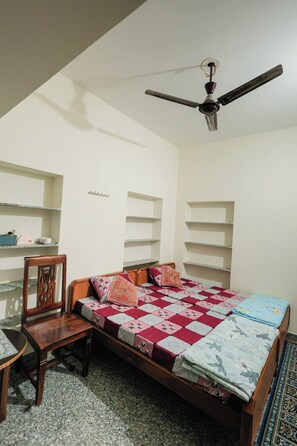 A bedroom with a bed, a chair, a ceiling fan, and built-in shelving. - The Jawai Niwas (Sumerpur)