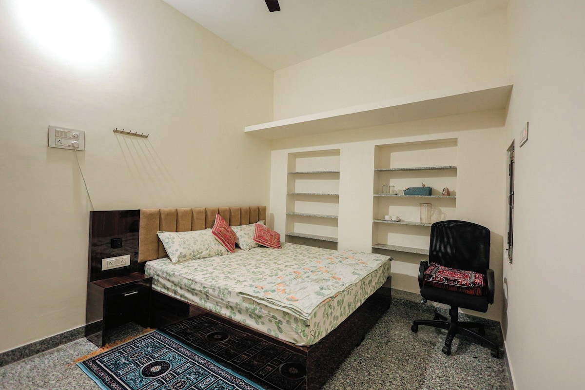 A bedroom with a bed, a desk, and a chair. There is a shelf unit against the wall and a power outlet.