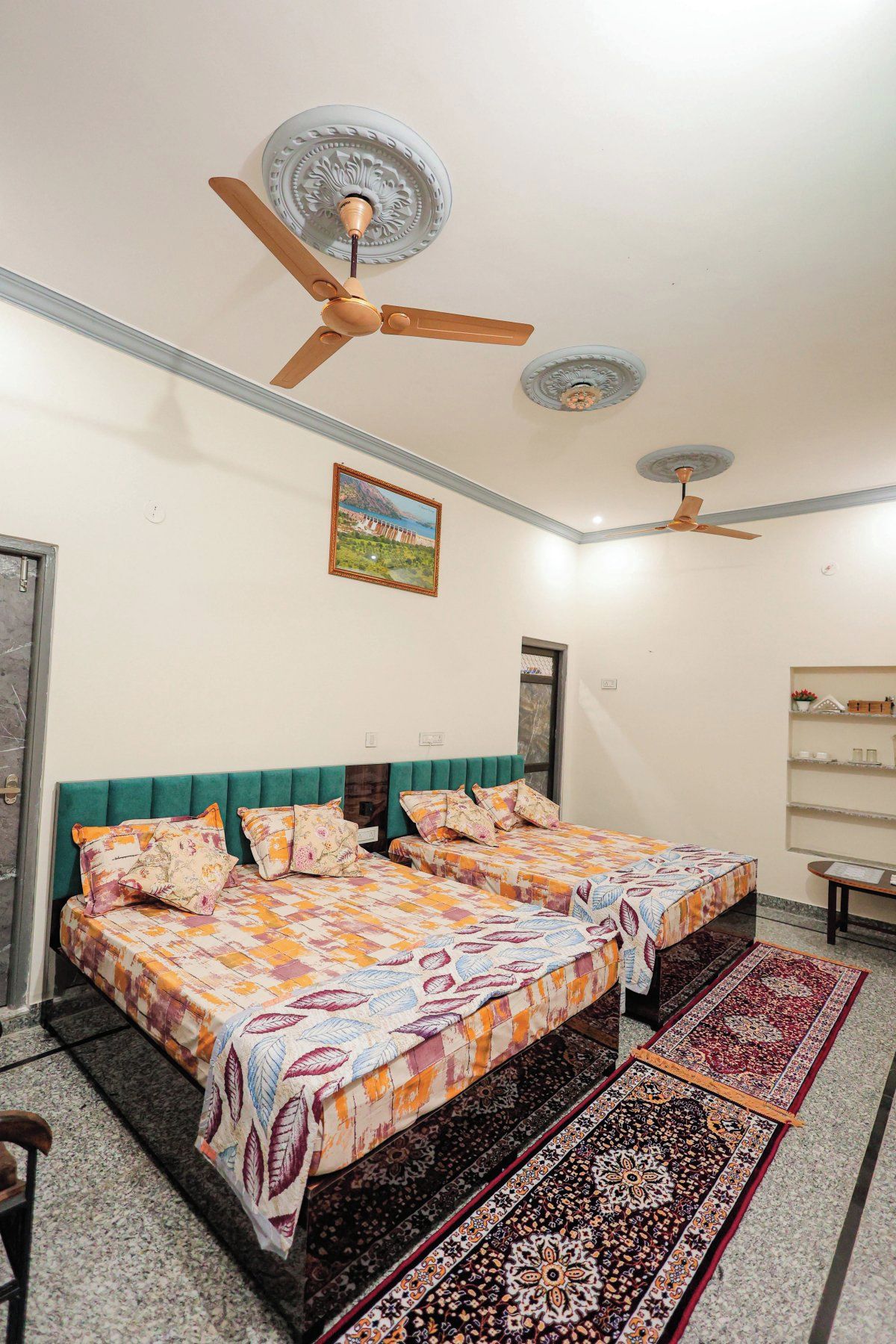 Un dormitorio con una cama doble, dos ventiladores de techo, un cuadro enmarcado y una mesita de noche con lámpara.