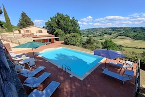 A swimming pool with lounge chairs and umbrellas, surrounded by a house and a view of rolling hills. - Villa in Riparbella with swimming pool (Riparbella)