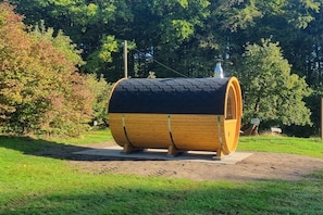 A wooden barrel-shaped structure with a black top, situated on a paved area with a grassy field and trees in the background. - Holiday home with 1 bedroom (Neuenkirchen)