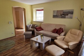 Ein Wohnzimmer mit Sofa, Sessel und Couchtisch. Es gibt ein Fenster mit Vorhang, eine Tür und ein Wandbild.