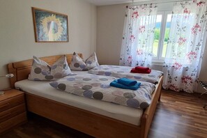 Ein Schlafzimmer mit einem hölzernen Bett, zwei Kissen, einer Decke, einem Nachttisch, einer Lampe, einem Fenster mit floralem Vorhang und einem Bild an der Wand.