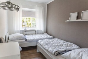 Ein Schlafzimmer mit zwei Betten, einem Heizkörper, einem Fenster mit Vorhängen und einem weißen Regal mit Bilderrahmen.