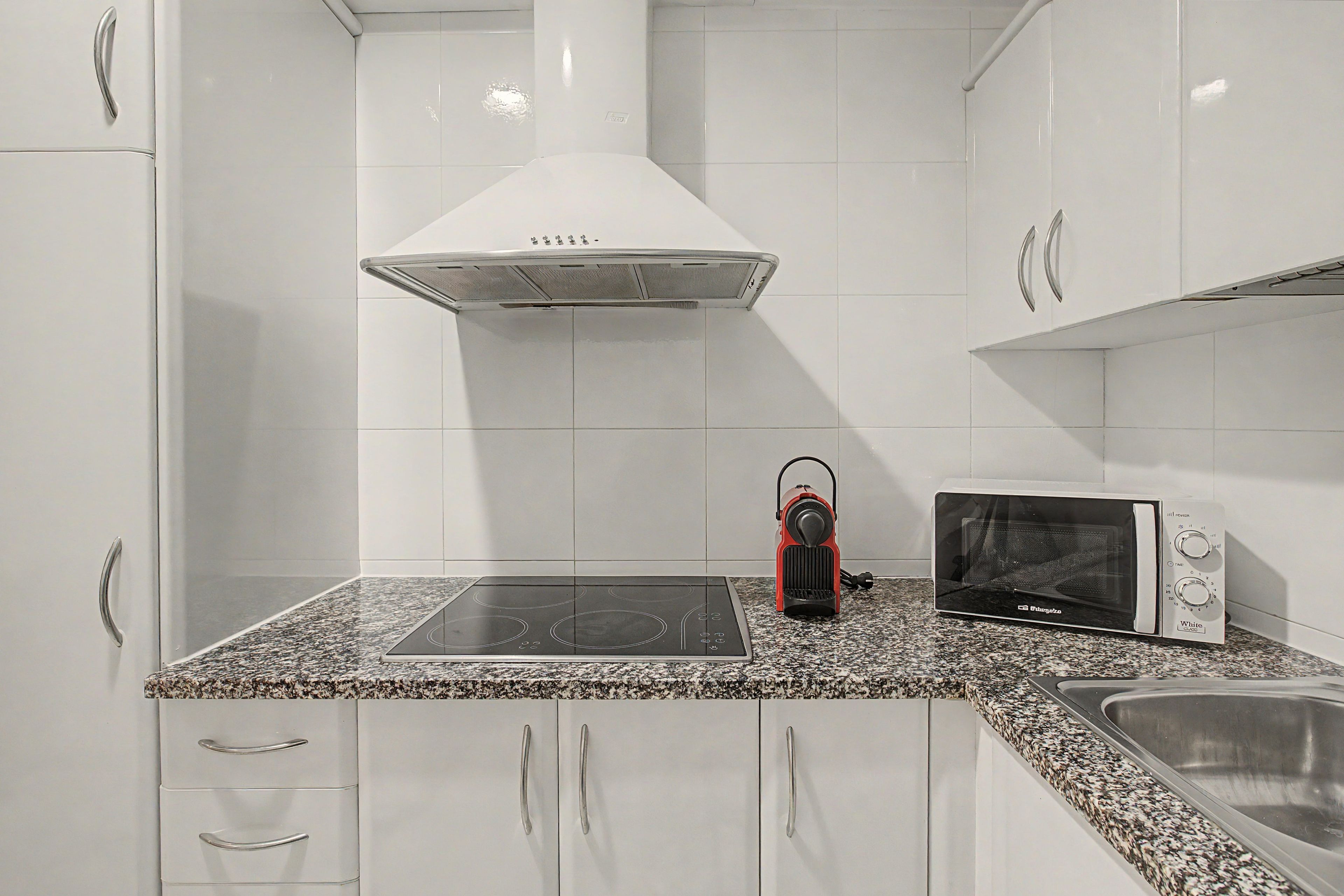 A kitchen with white cabinets, a black induction cooktop, a red kettle, and a white microwave.
