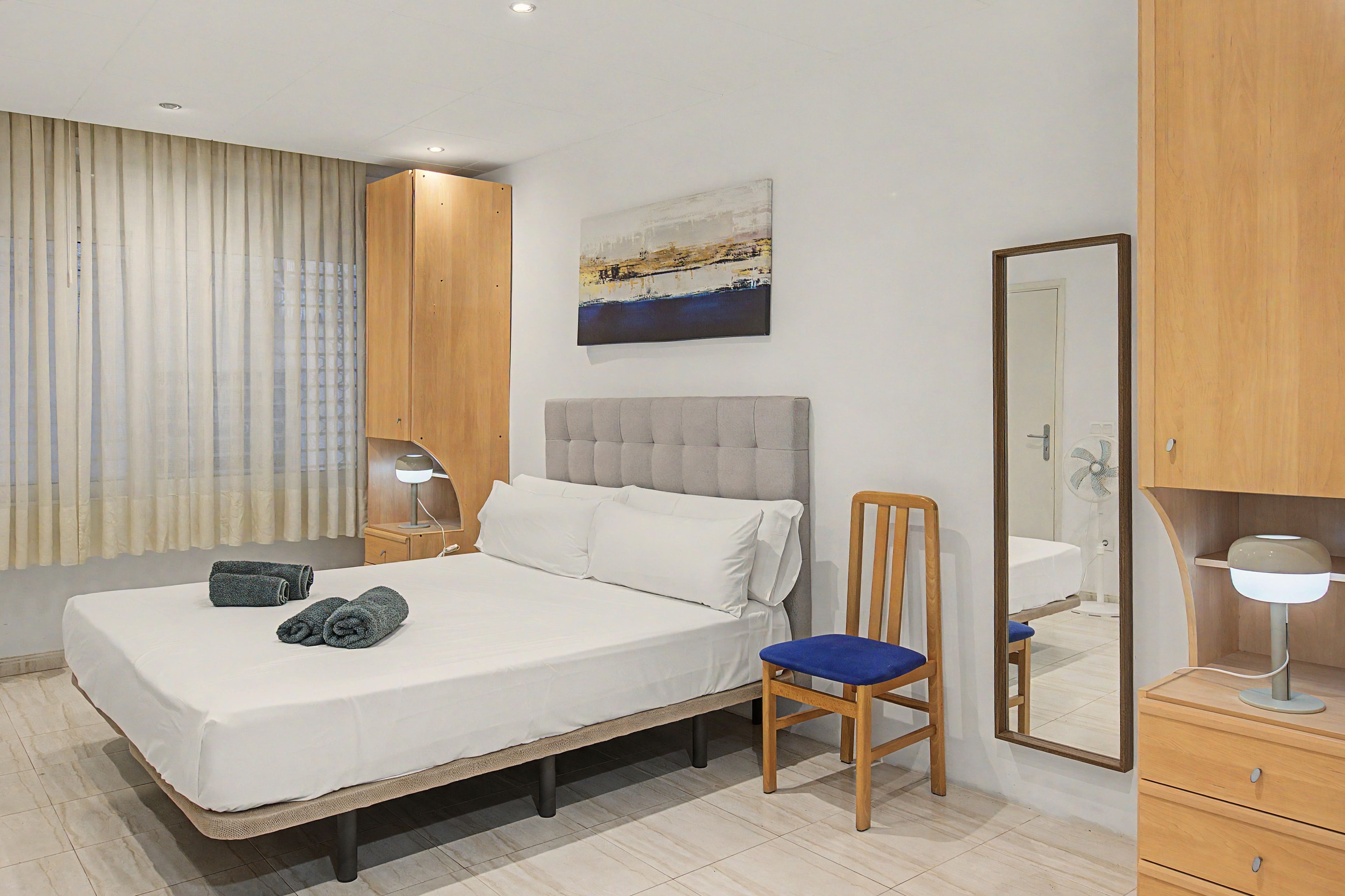 Habitación de hotel con cama, una silla, un escritorio con lámpara, espejo y un cuadro en la pared.