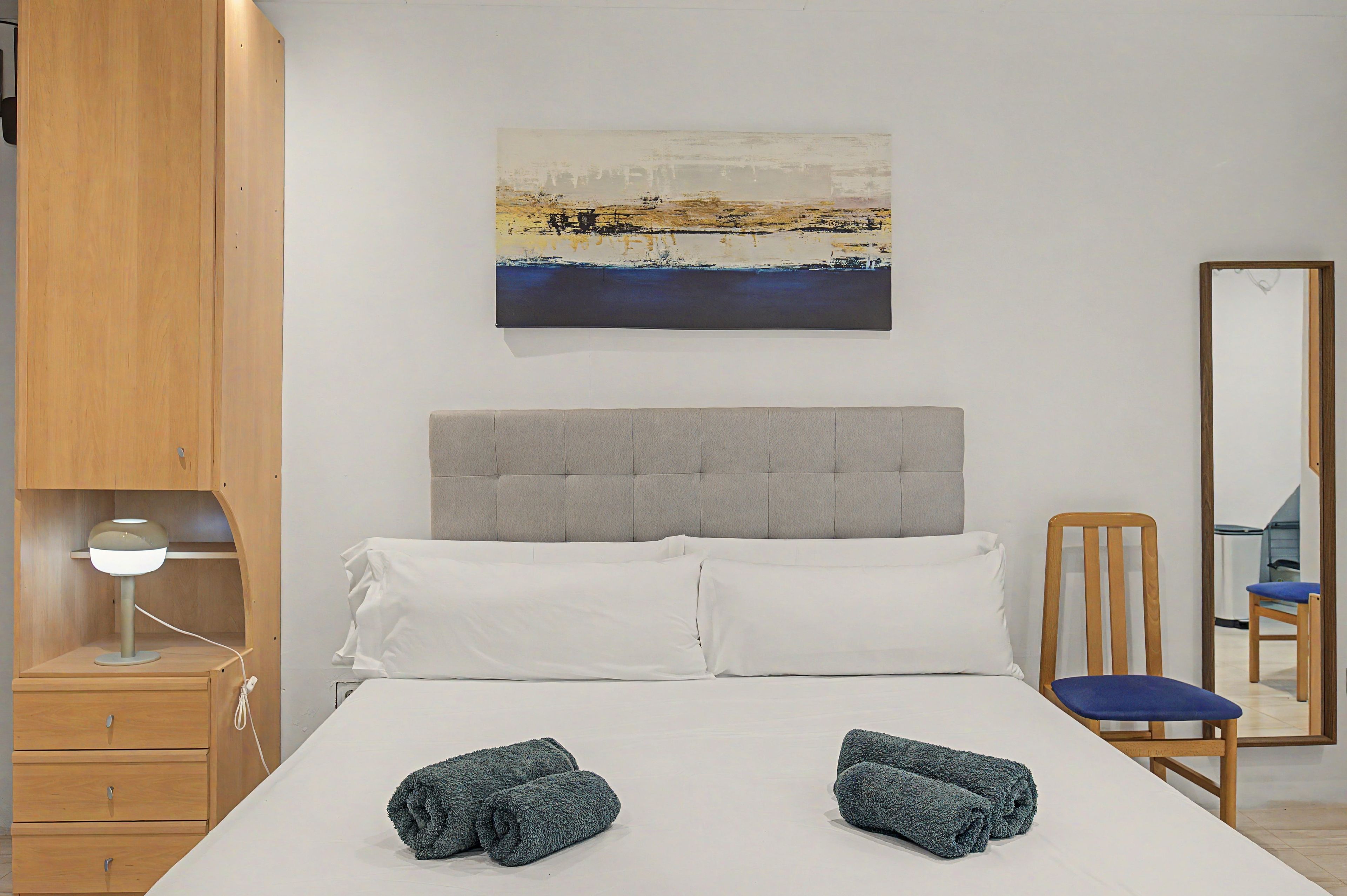 A hotel room with a bed, a bedside table with a lamp, a chair, a painting on the wall, and a mirror.