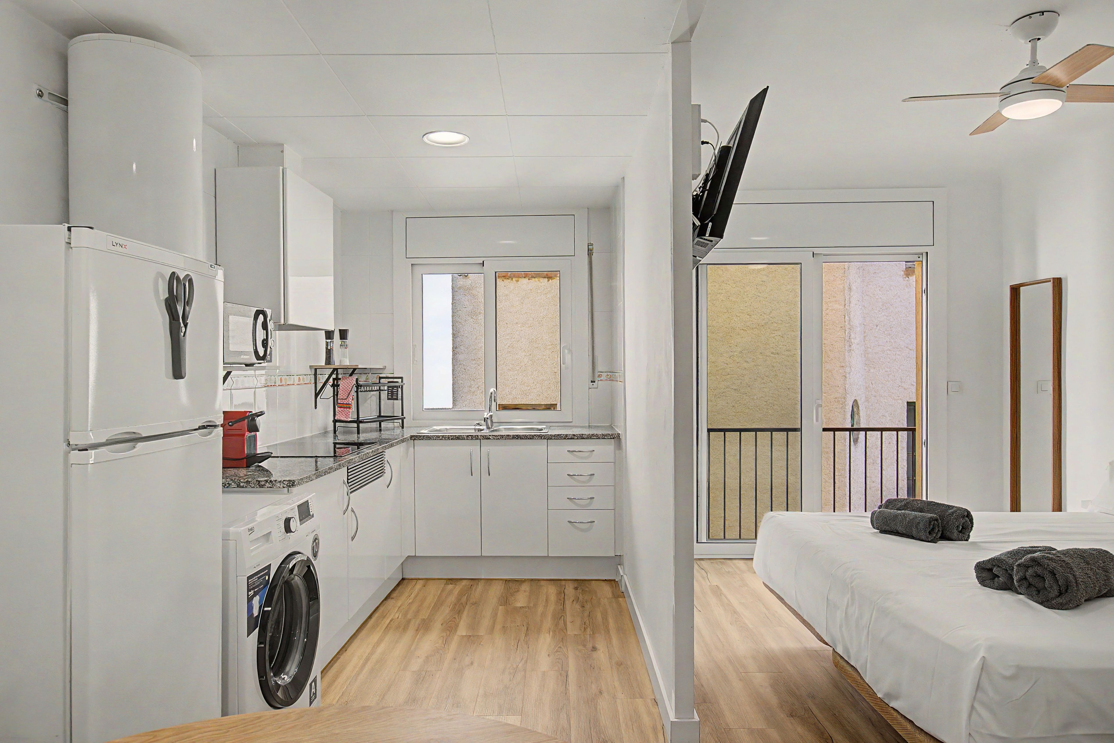 A compact hotel room with a kitchenette, a washing machine, a bed with towels, and a balcony with a view.