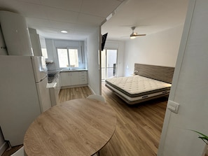 Een kleine kamer met een bed, een tafel, een kitchenette en een plafondventilator.