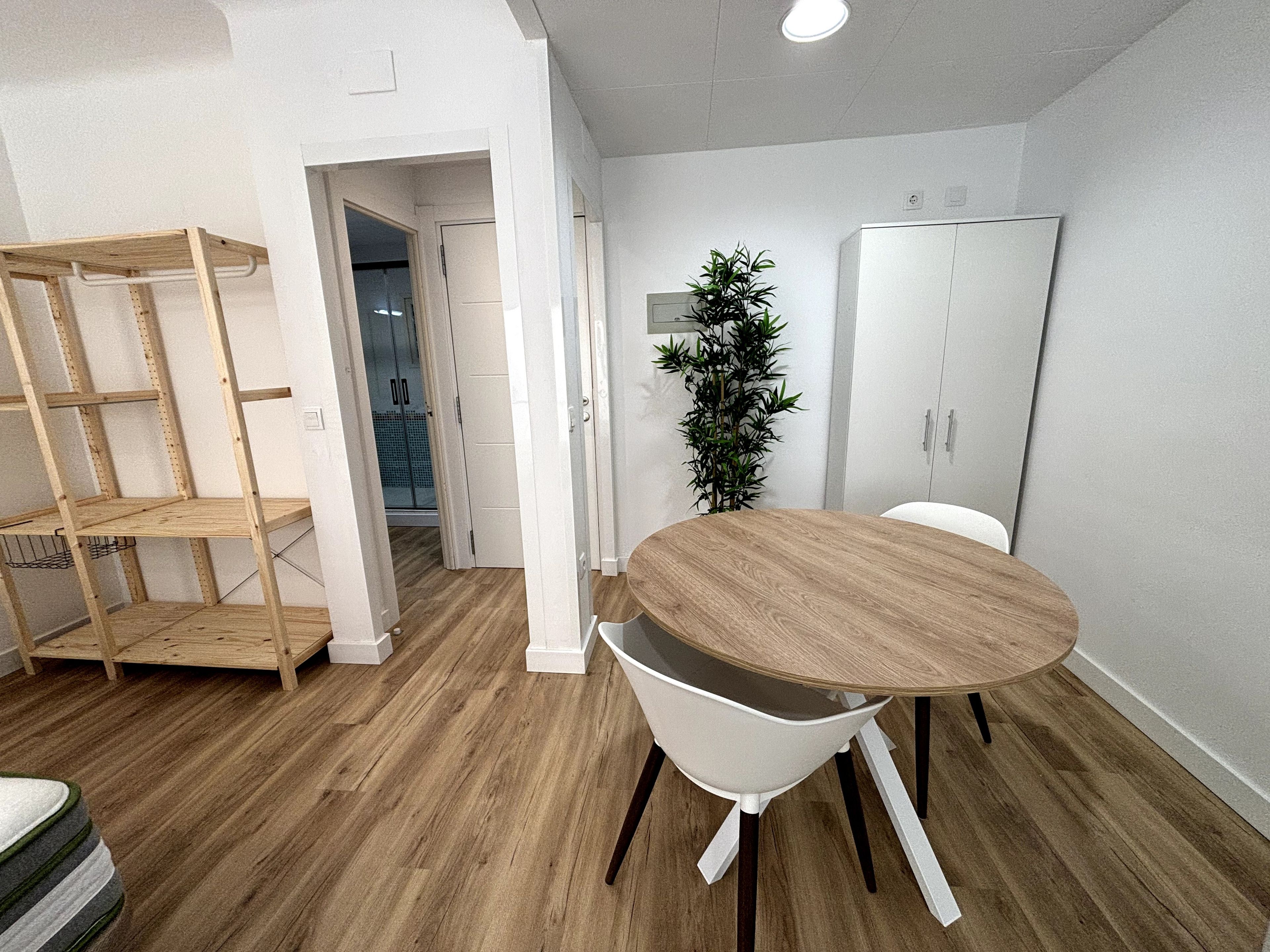 A room with a round wooden table, white chairs, a plant, and a wardrobe.
