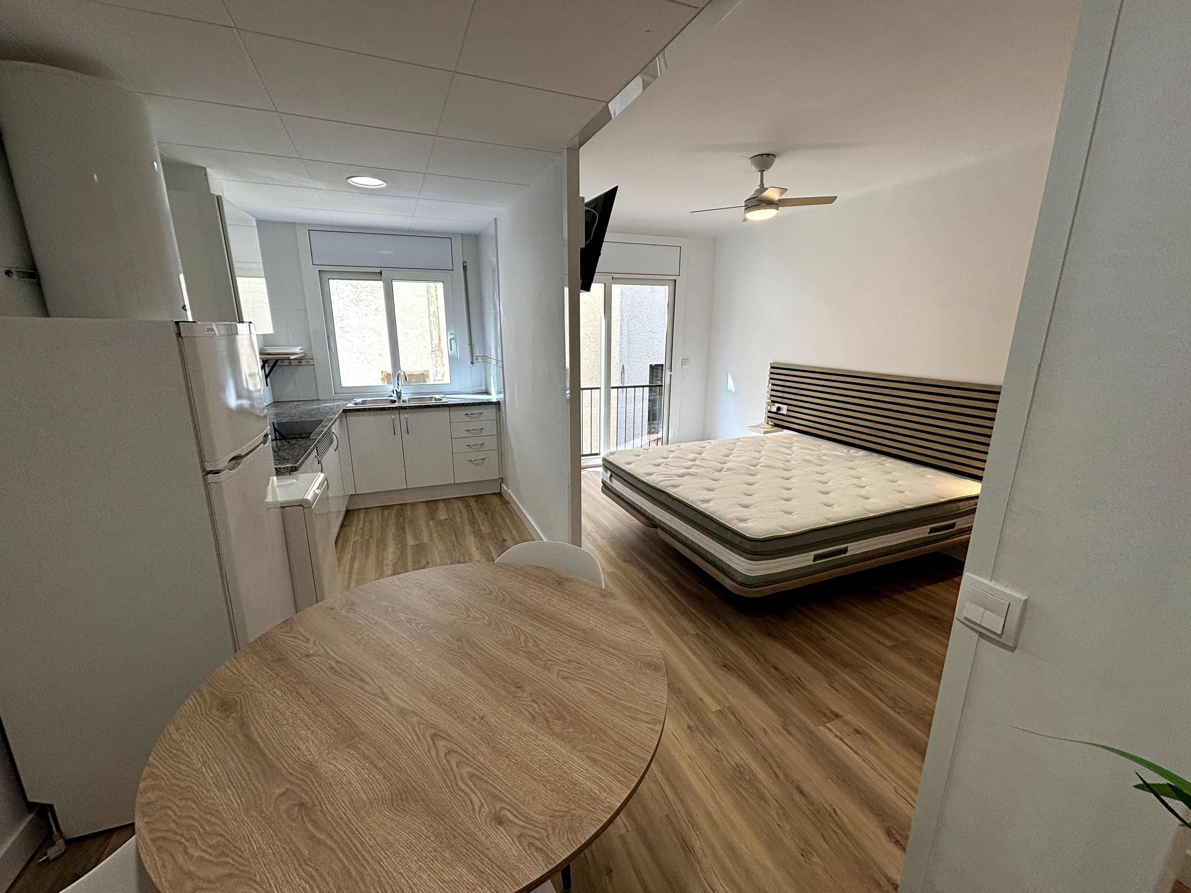 A compact room with a bed, a table, a kitchenette, and a ceiling fan.