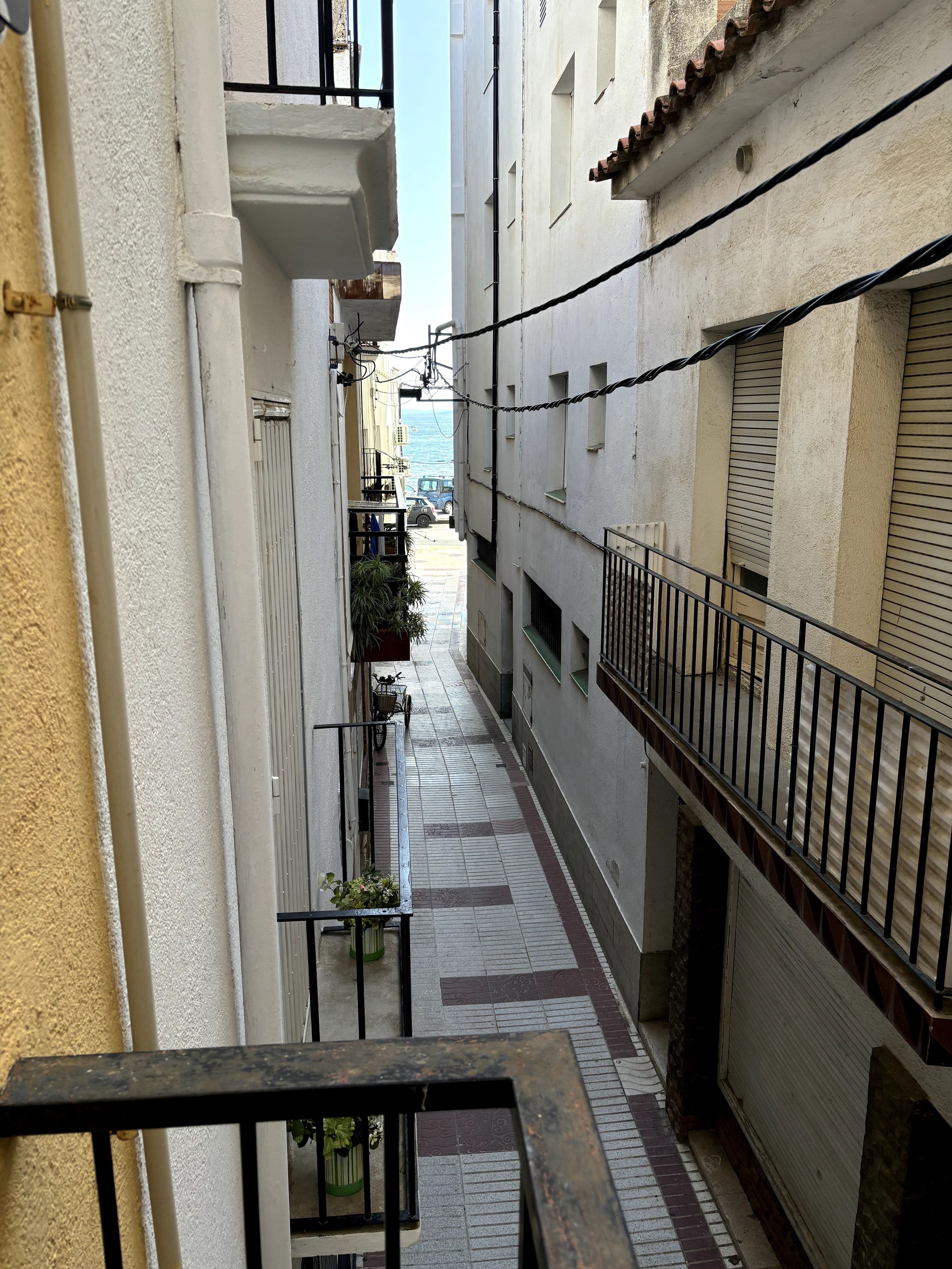 A narrow urban alleyway with buildings on either side, a bicycle, and a distant view of the sea.