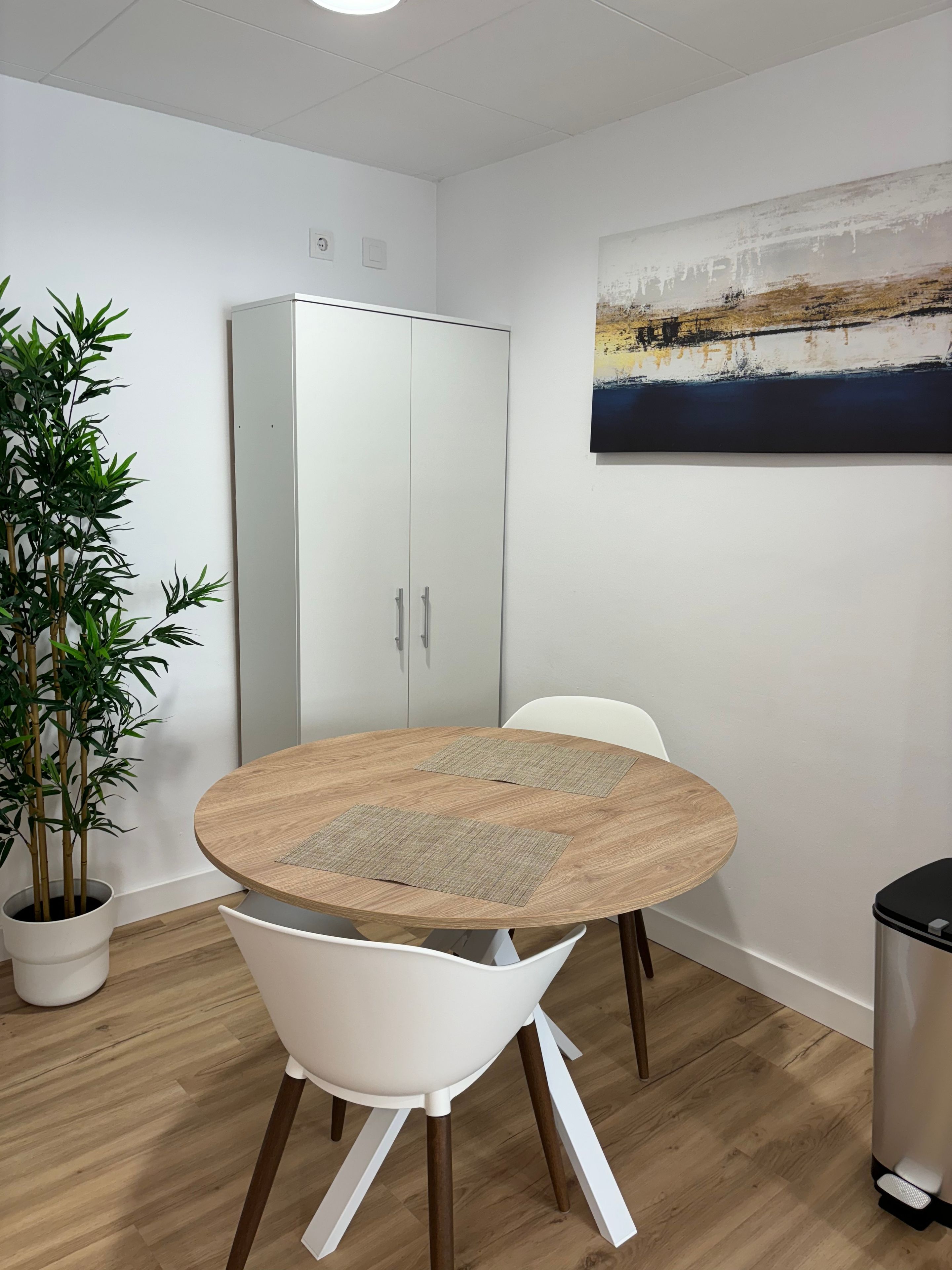 Una habitación con una mesa redonda de madera, sillas blancas, una planta en maceta, un armario y un cuadro en la pared.