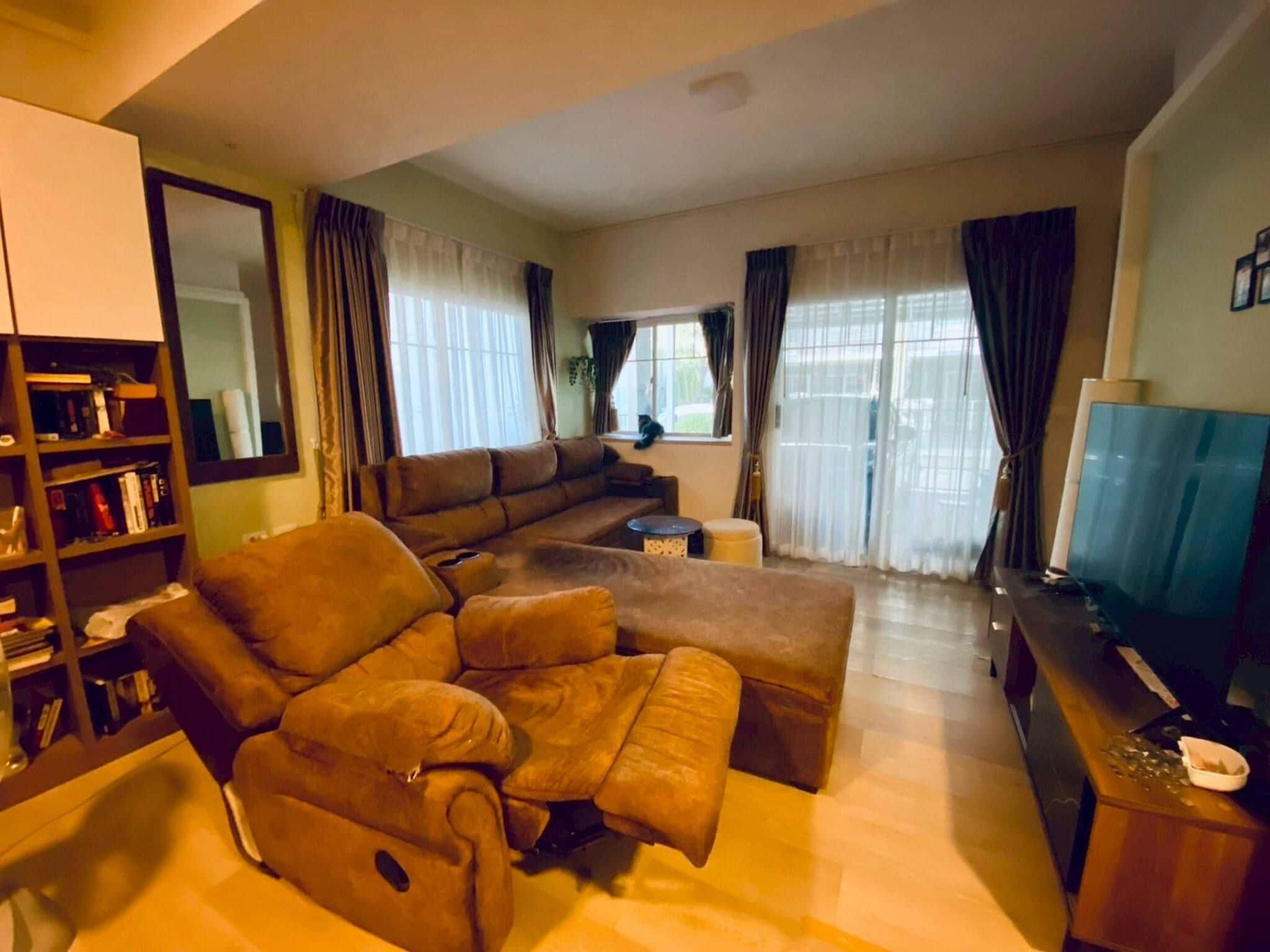 A living room with a sectional sofa, a television, a bookshelf, and a large window with curtains.