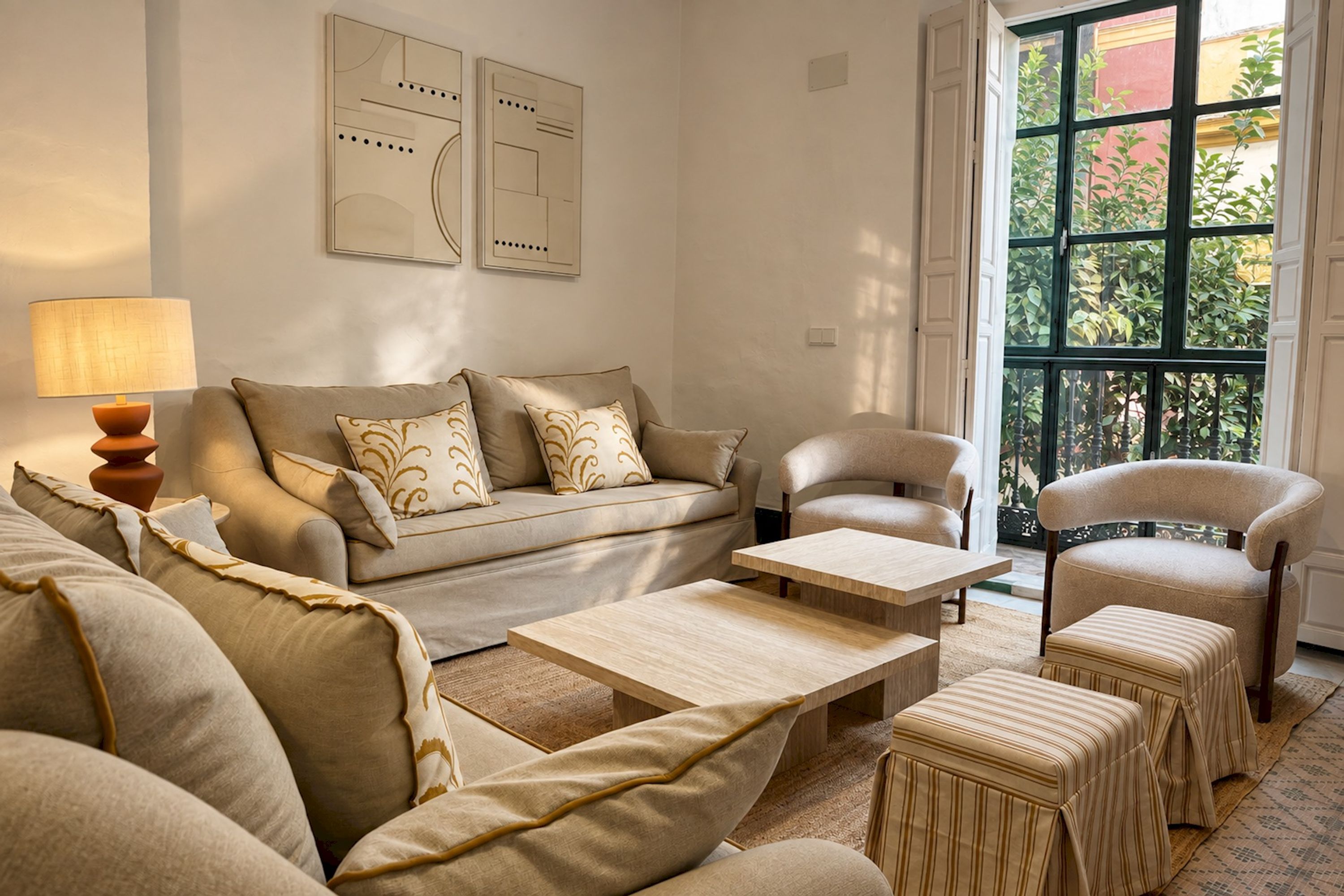 A living room with a sofa set, a coffee table, a side table with a lamp, and a view of greenery through the window.