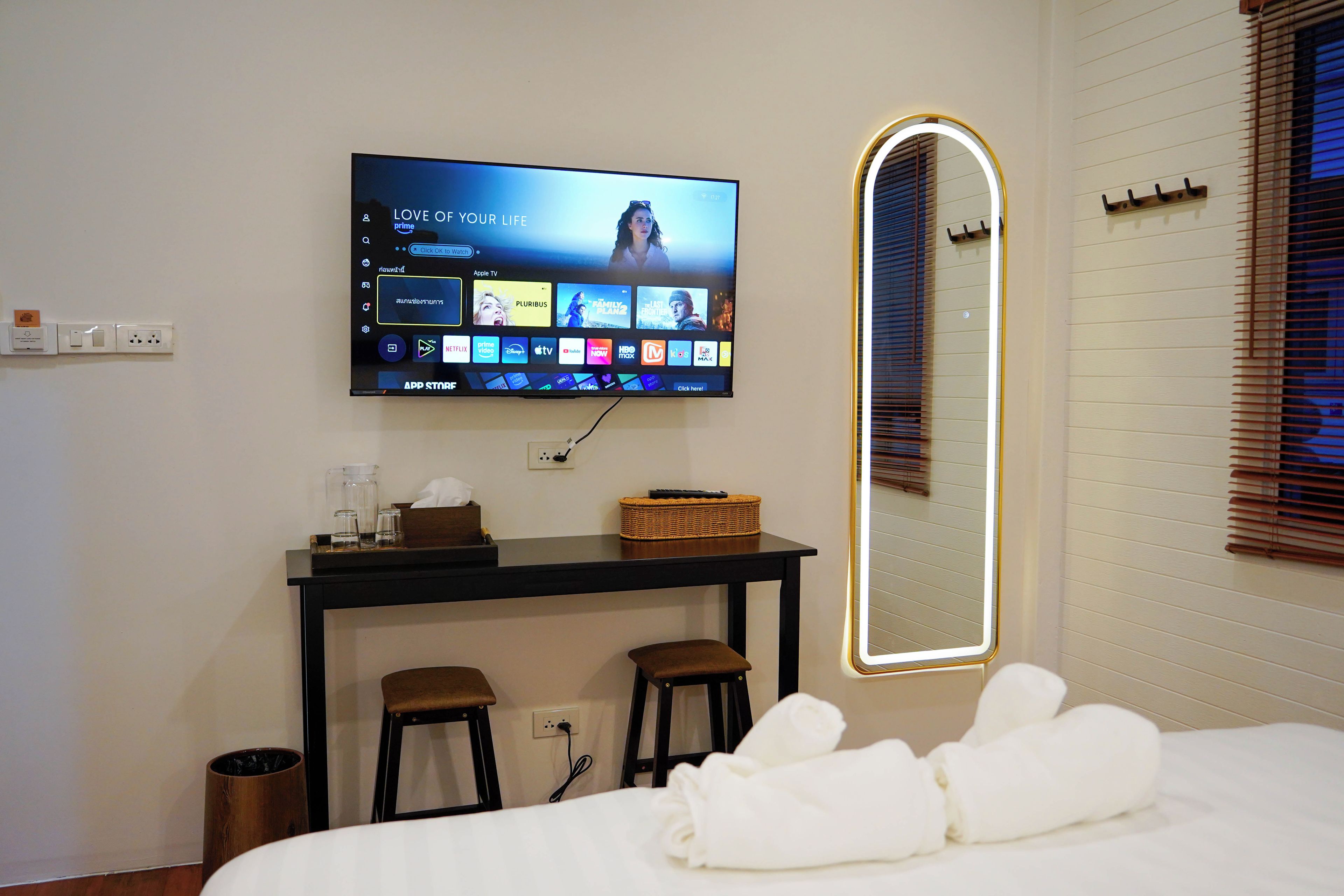 Habitación de hotel con televisor de pantalla plana montado en la pared que muestra varios servicios de streaming, una mesita con una cesta y vasos, una cama con toallas blancas y un espejo con marco iluminado.