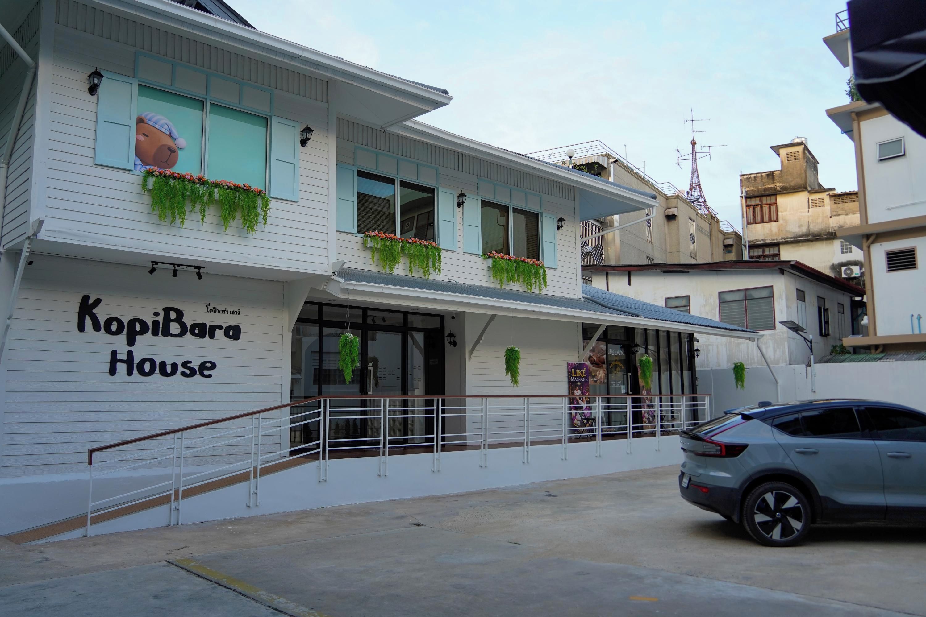 Un edificio de dos pisos con un letrero que dice 'KopiBara House'. Hay macetas con flores en las ventanas, un balcón con barandilla y un coche estacionado adelante.