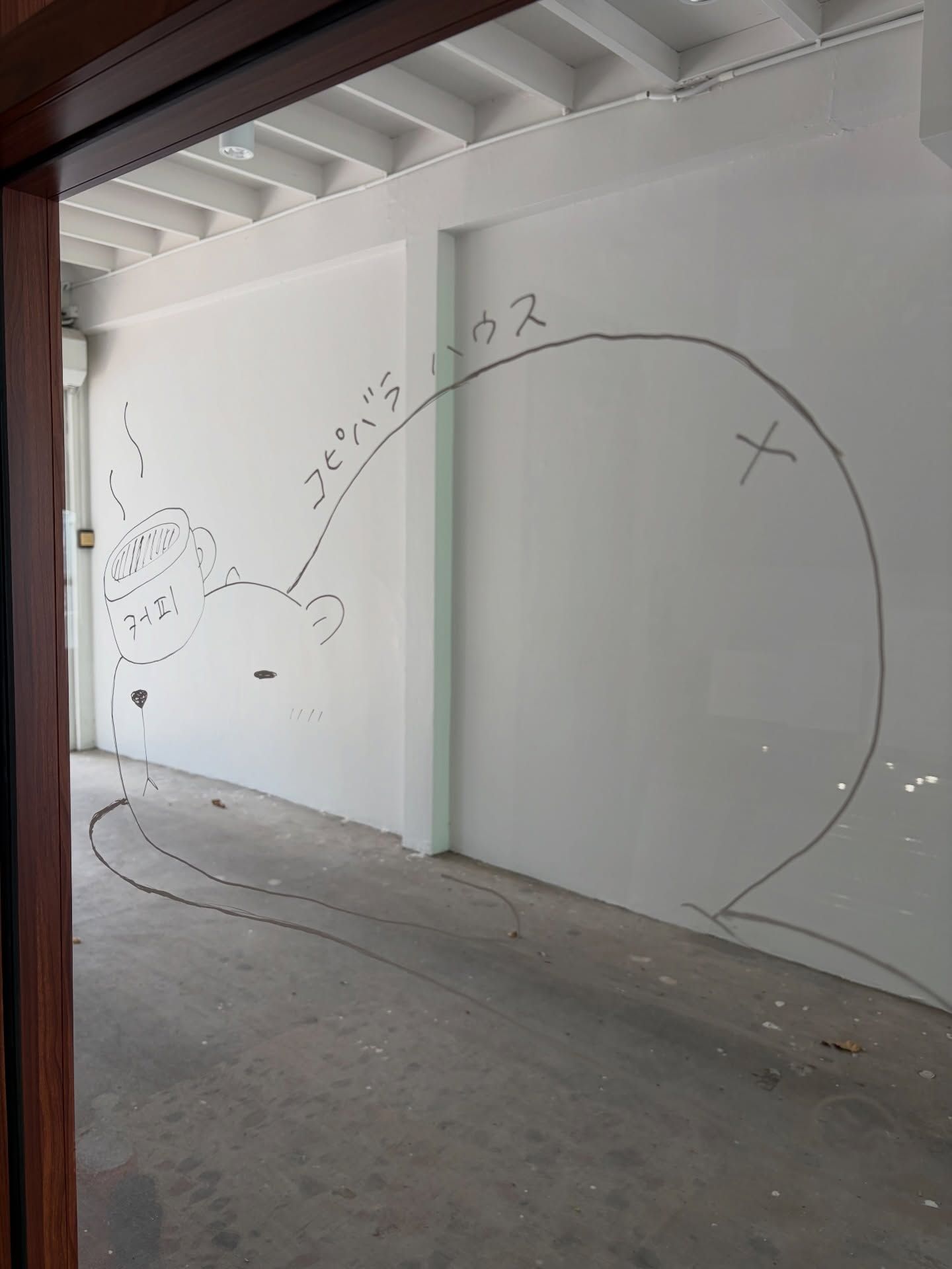 A room with white walls, a concrete floor, and a large window. There is a drawing of a bear and a cup on the wall.
