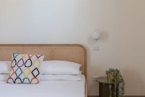 Un lit avec une tête de lit en osier, un coussin coloré, du linge de lit blanc, une lampe de chevet et une plante en pot sur une table de chevet.
