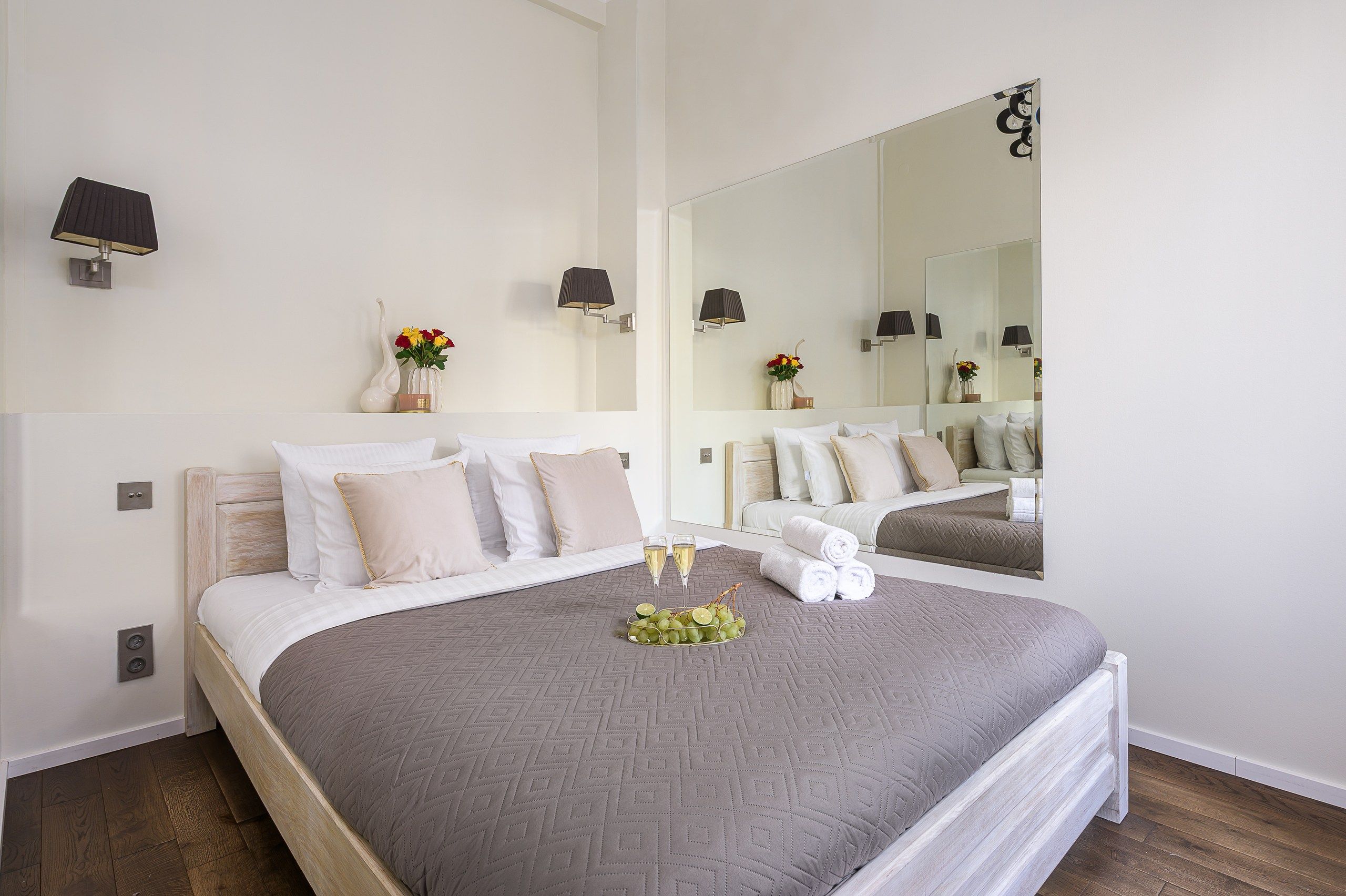 A hotel room with a large bed, a mirror, wall-mounted lamps, and a vase with flowers.