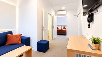 A hotel room with a sofa, a small table, a bedside table, a bed, a wall-mounted phone, and an air conditioning unit.