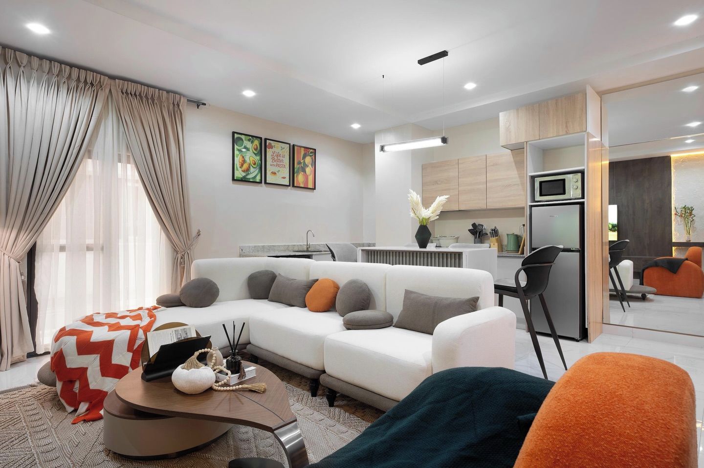 Una sala de estar moderna con un sofá, una mesa de centro y un reposapiés. Hay una zona de cocina con microondas y refrigerador.