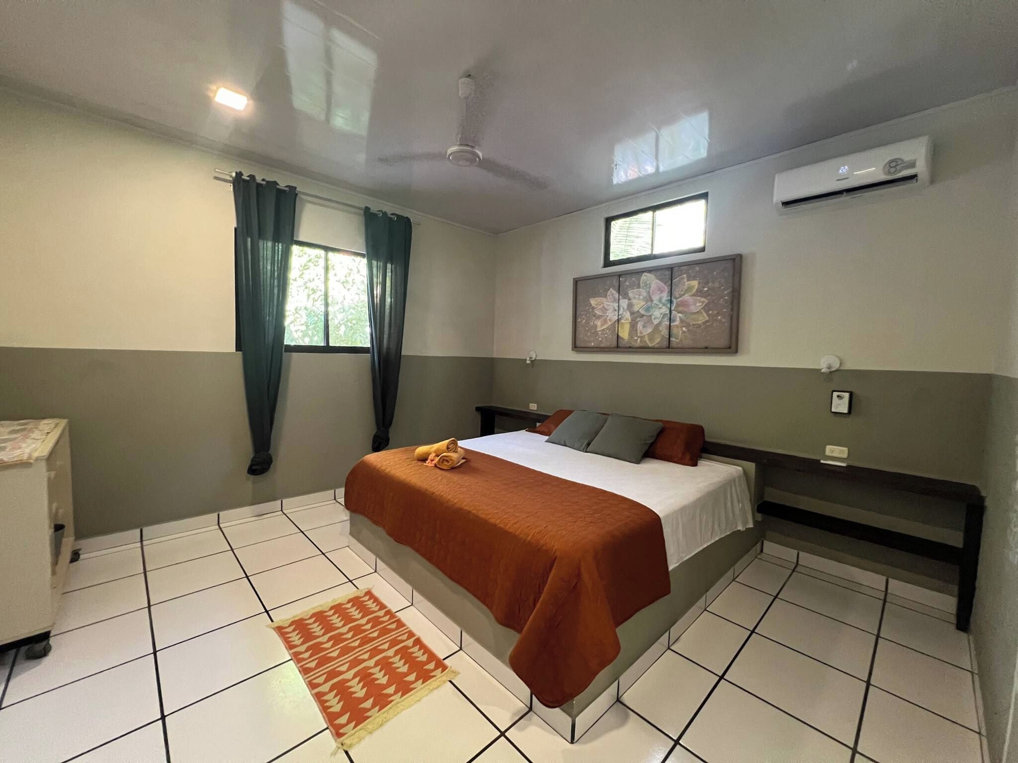 Habitación de hotel con cama, ventana con cortinas, ventilador de techo, aire acondicionado y suelo de baldosas.