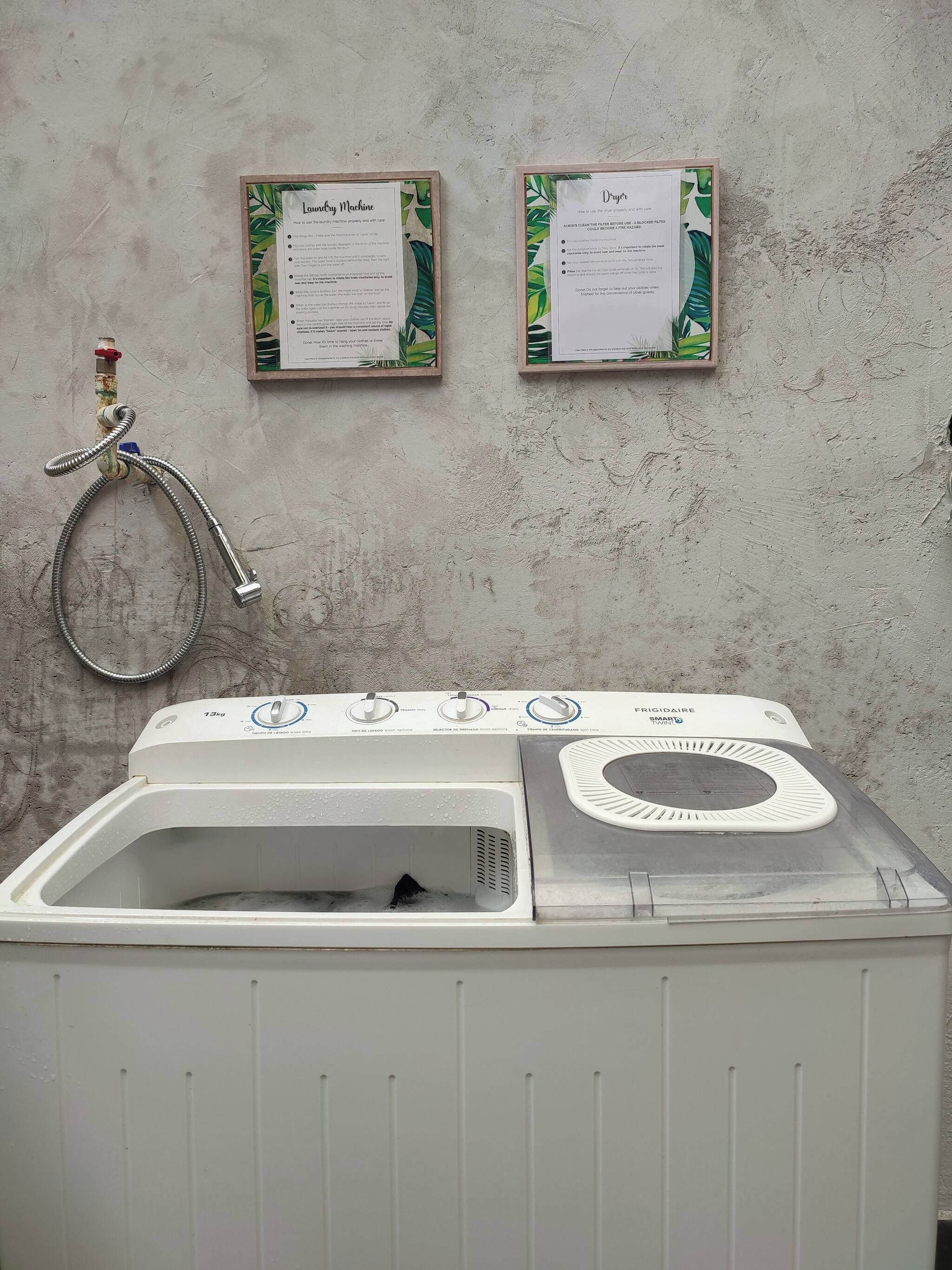 A front-loading washing machine with a control panel, a hose, and two informational posters on the wall.