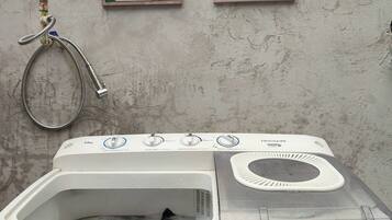 A front-loading washing machine with a control panel, a hose, and two informational posters on the wall.