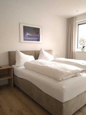 Une chambre d’hôtel moderne avec un grand lit, une table de chevet, une commode, une fenêtre avec des rideaux et un tableau encadré au mur.