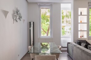 A modern living room with a glass dining table, a sofa, and a window with a view of greenery. - iFlat | Sleek & Sunlit Apartment near the Vatican (Rome)