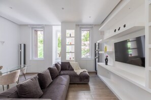 A modern living room with a sectional sofa, a glass dining table, and a large flat-screen TV mounted on a white wall. - iFlat | Sleek & Sunlit Apartment near the Vatican (Rome)