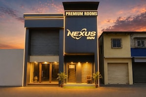 Un hotel con un cartel que indica 'Nexus Inn' y 'Habitaciones Premium', mostrando un edificio moderno con una entrada destacada y una escalera que conduce a la recepción.
