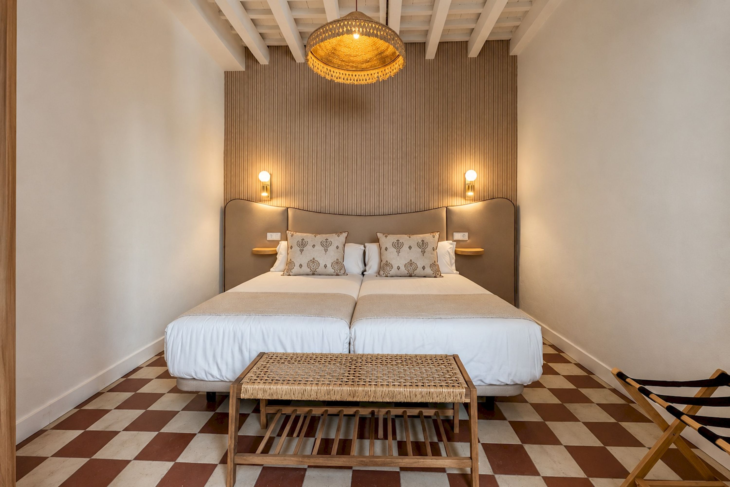 A bedroom with a large bed, two bedside lamps, a woven bench, and a checkered floor.