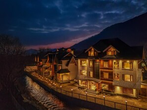 A row of multi-story buildings with balconies, a river in the foreground, and mountains in the background. - The Harmukh Hills Pahalgam (Pahalgam)