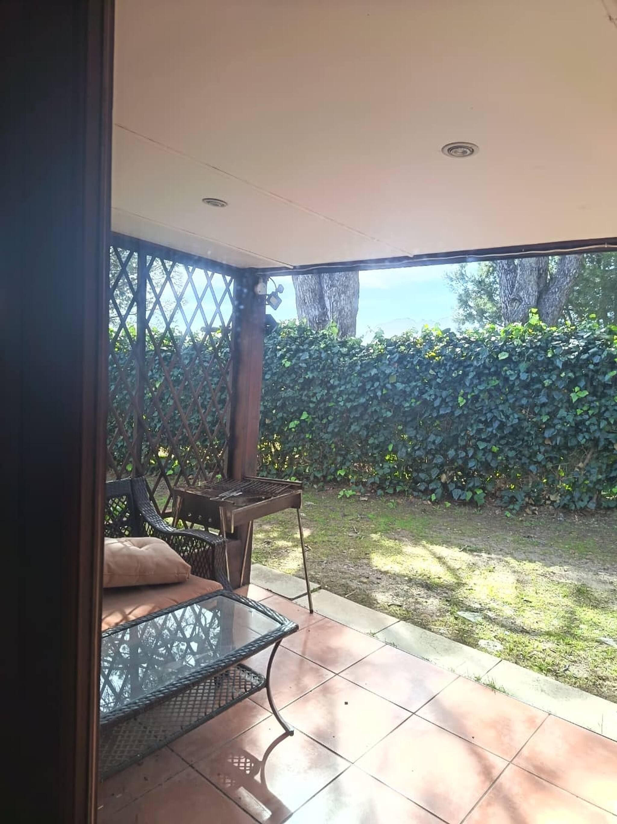 A covered patio with a grill, a table with chairs, and a view of a hedge and trees.