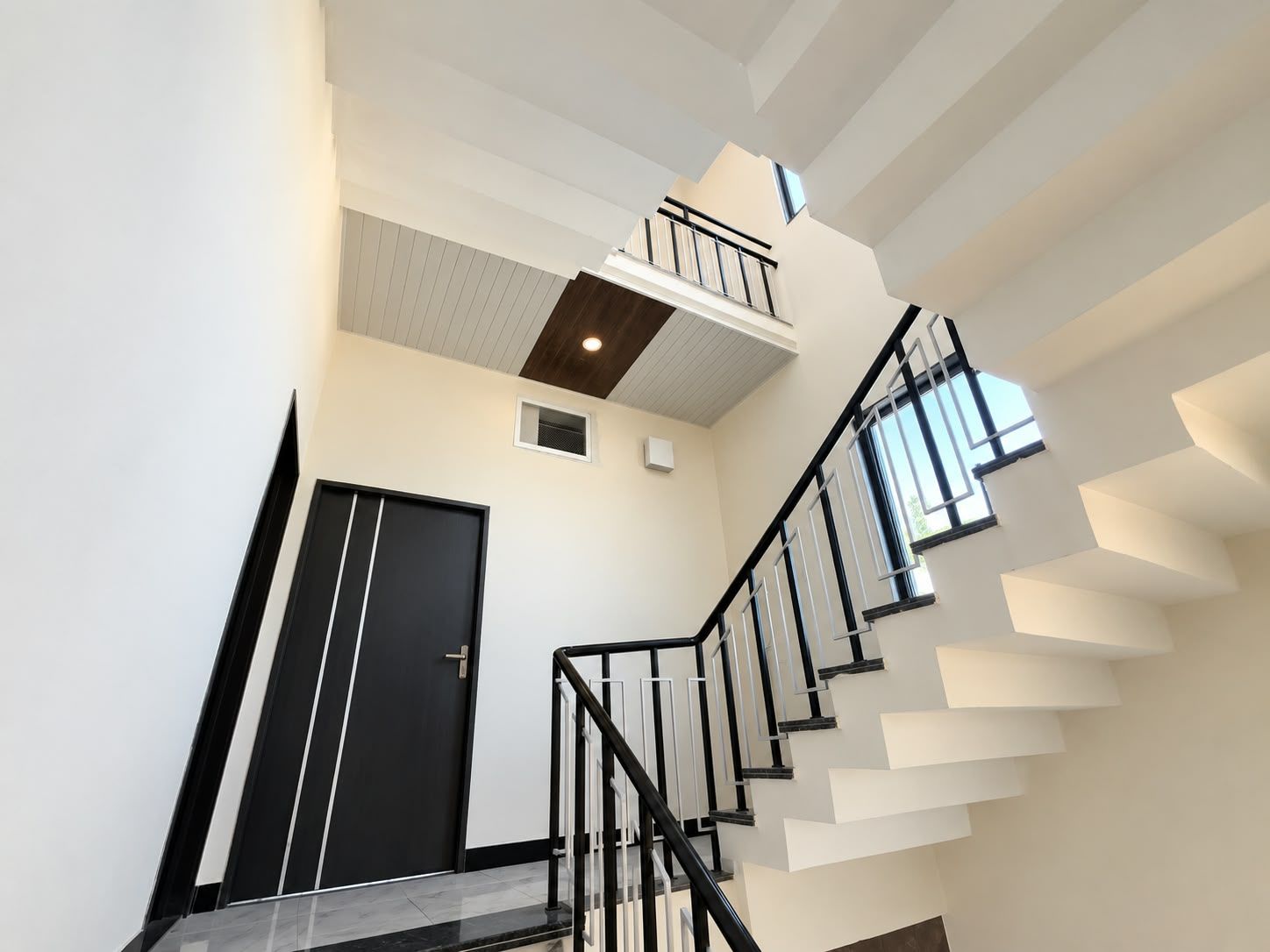 Una escalera moderna con barandilla negra y paredes blancas, que conduce a una plataforma con un ventanal pequeño.