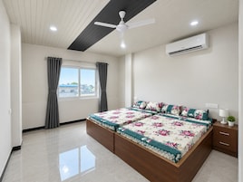 Un dormitorio con cama, ventilador de techo, equipo de aire acondicionado, ventana con cortinas y vistas a edificios.