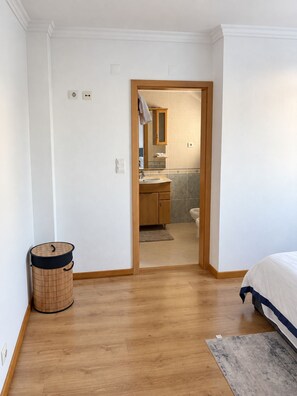 Ein Schlafzimmer mit Holzboden, einem Bett mit weißer und blauer Bettdecke, einer kleinen Mülltonne und einer Tür zu einem Badezimmer mit Waschbecken und Spiegel.