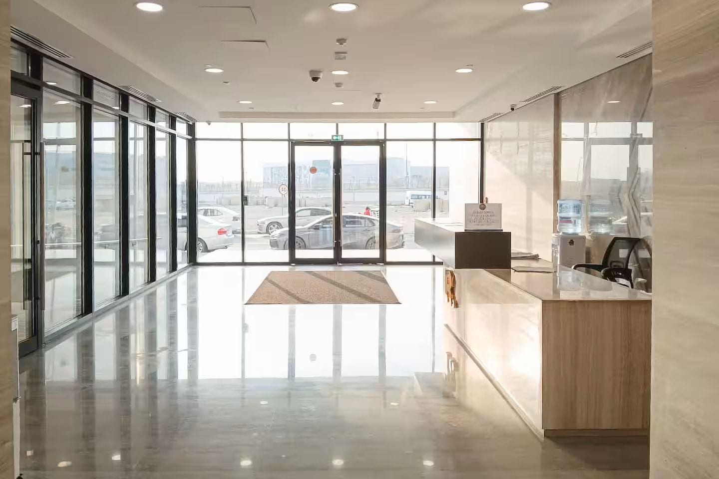 Un área de recepción moderna con entrada por puerta de vidrio, un mostrador de recepción y vista al estacionamiento.