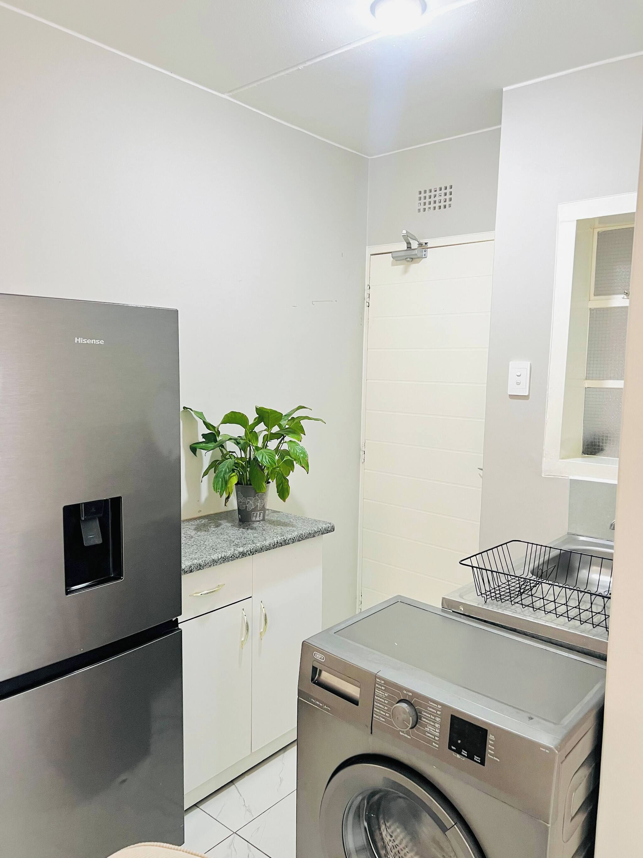 A compact kitchen with a stainless steel refrigerator, a washing machine, and a potted plant on the countertop.