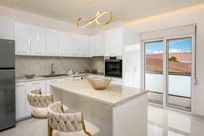 A modern kitchen with white cabinetry, a central island, and a view of a red-roofed building outside. - Annamaria's Apartment (Rodos)