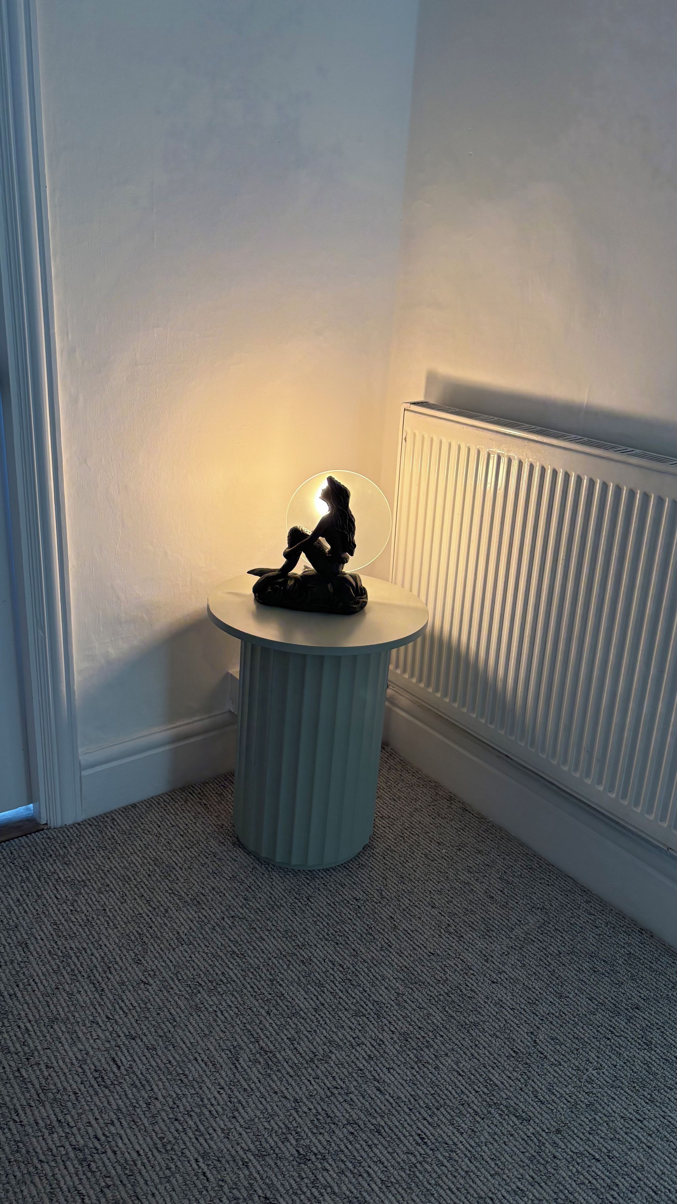 A corner room with a radiator, a small round table with a sculpture, and a carpeted floor.