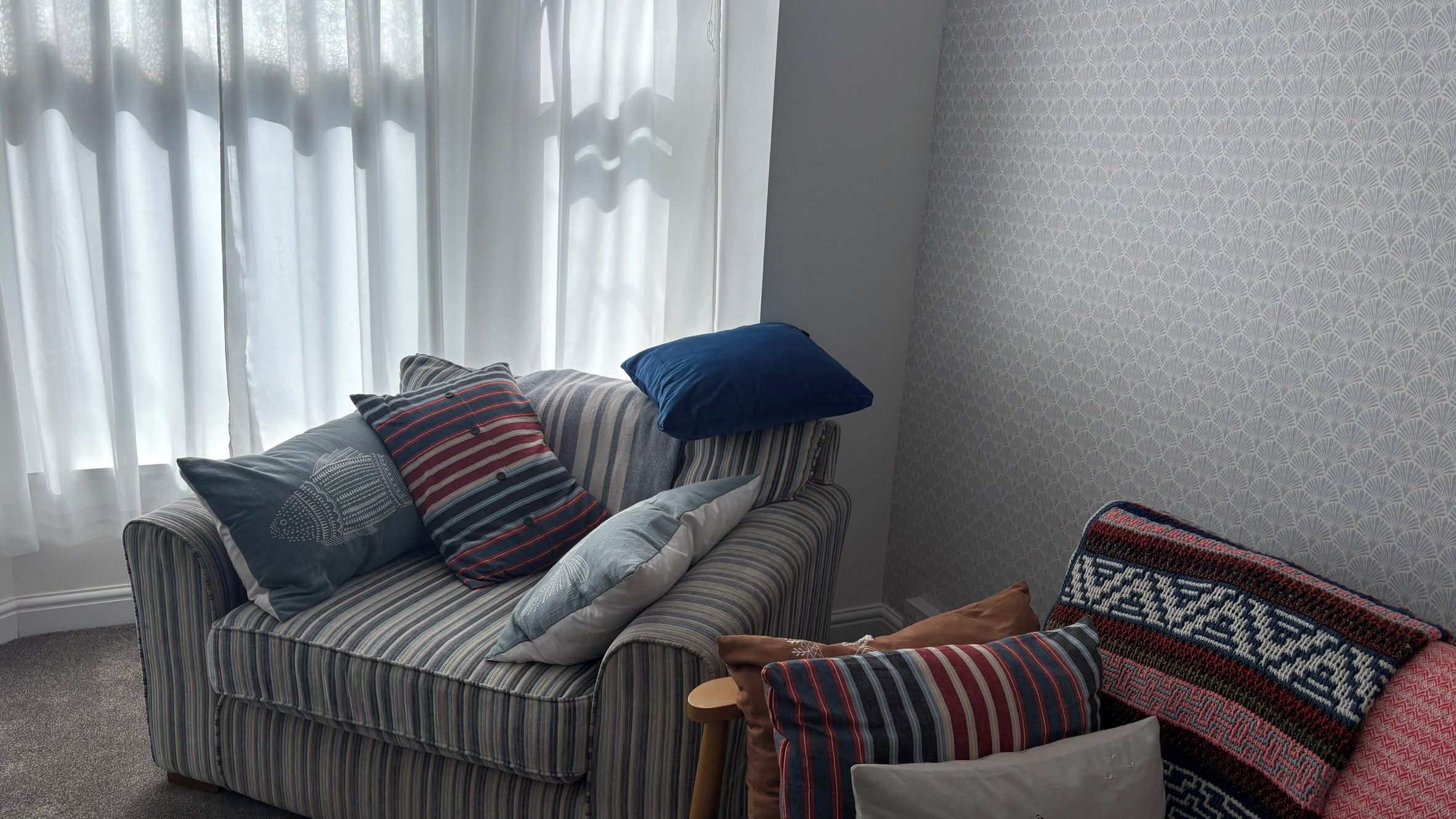 Ein gestreiftes Sofa mit vielen Kissen, ein blaues Kissen und eine gemusterte Decke in einem Raum mit Wandmuster und transparenten Vorhängen.