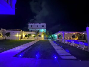 A nighttime view of a hotel courtyard with a pool, lounge chairs, and a building with illuminated arches. - Sunset Apartments Bagamoyo (Bagamoyo)
