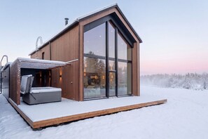 Una cabaña moderna con un amplio ventanal, una terraza de madera y una zona de sauna.
