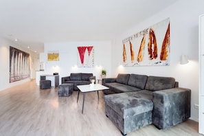 Uma sala de estar moderna com um sofá seccional cinza, uma mesinha branca e um banquinho cinza. Há duas pinturas na parede, uma com cores vermelhas e brancas e outra com barcos à vela.